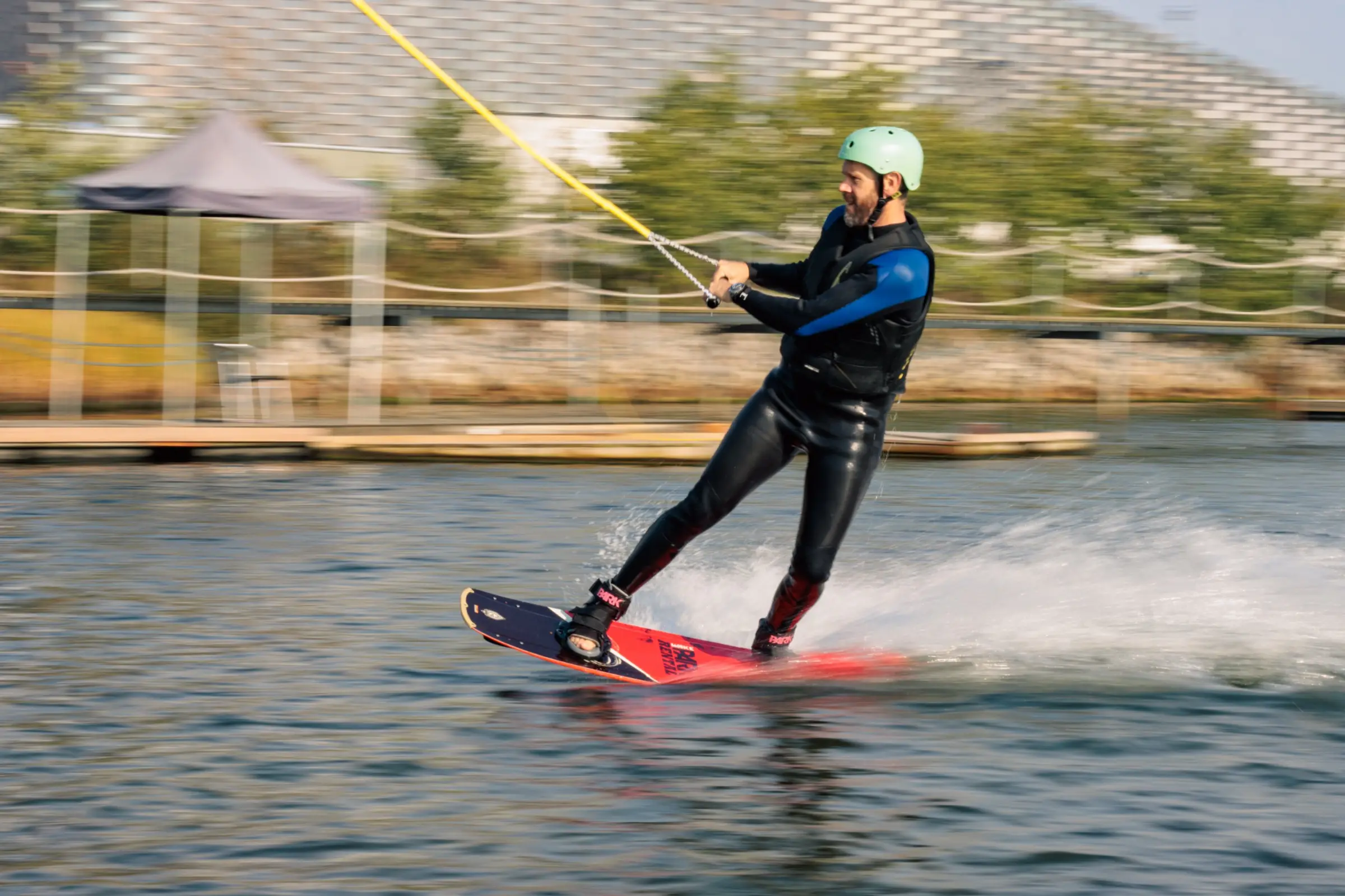A rider in a wetsuit and helmet on a rental wakeboard, passing the dock with Copenhagen behind them